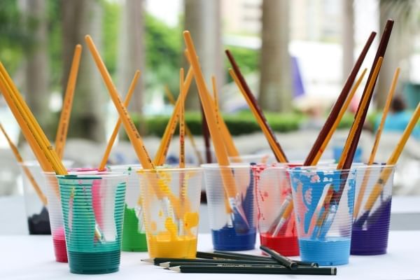 Eight plastic transparent cups filled with multicolor paints. Paintbrushes stick out of the cups. Several pencils lie on the table beside them. 