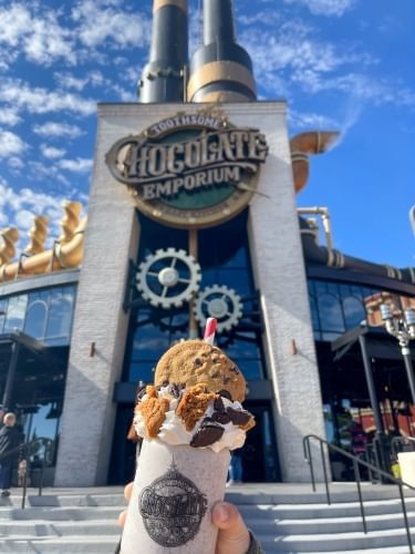 A tall entryway with a smokestack and a sign that reads Toothsome Chocolate Emporium, slightly out of focus with a hand holding an enormous milkshake topped with cookie pieces and a straw in the foreground. Toothsome Chocolate Emporium is a great CityWalk restaurant.