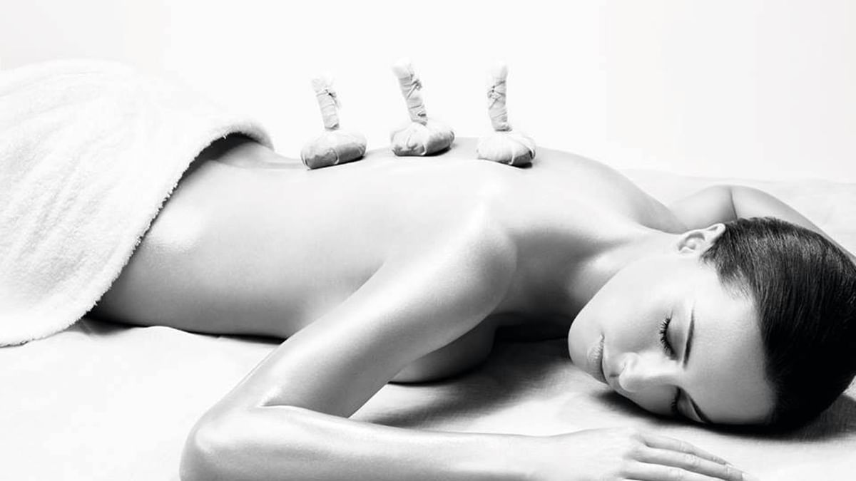 Femme allongée sur un lit de massage avec des herbes sur son dos, yeux fermés.