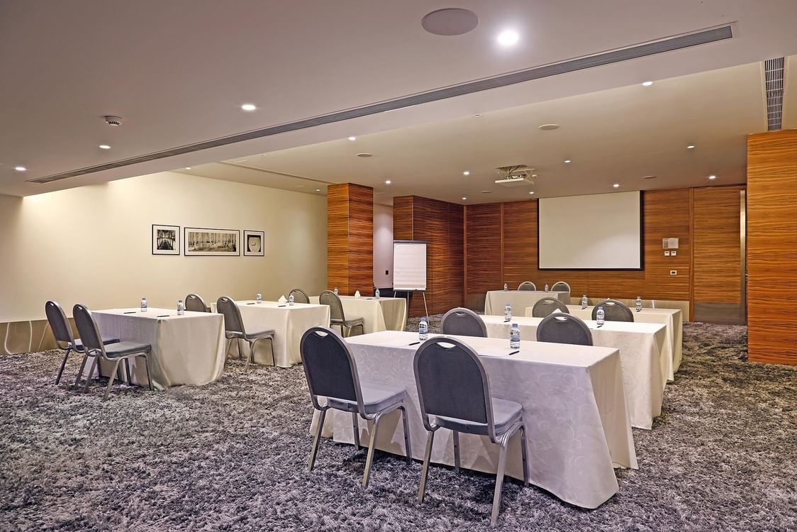 Grey chairs by tables under a projector screen near wood-paneled pillars in Emerald Room at Warwick Stone 55 - Beirut