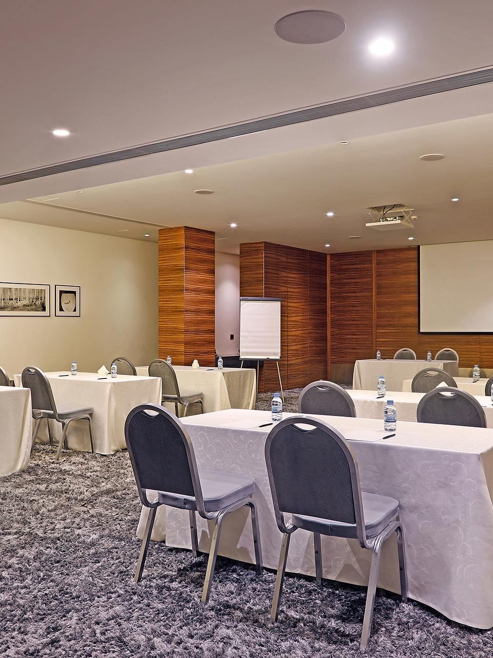 Grey chairs by tables under a projector screen near wood-paneled pillars in Emerald Room at Warwick Stone 55 - Beirut