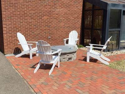 White Adirondack chairs around a round stone fire pit on a red brick terrace against a brick wall at The Simsbury Inn