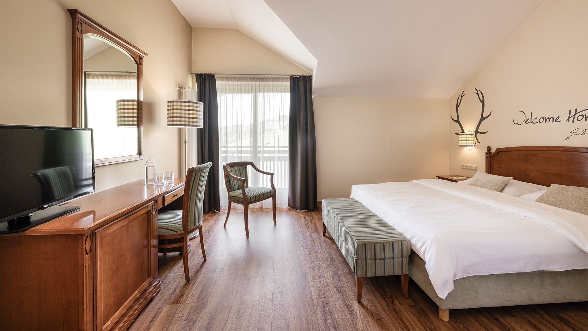 Geräumiges Hotelzimmer mit Doppelbett, Sitzbank, Schreibtisch, Fernseher und Balkon mit Vorhängen.