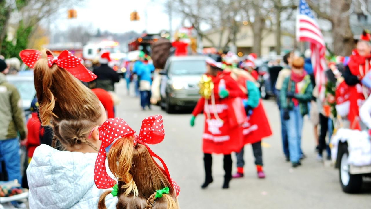 Children with festive red bows in their hair watch a parade along a street lined with spectators.