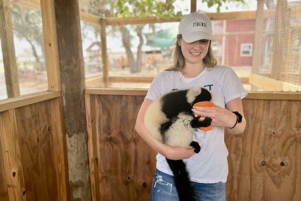 A young woman in a baseball hat holds a lemur in one arm and an orange cup of fruit in the other.