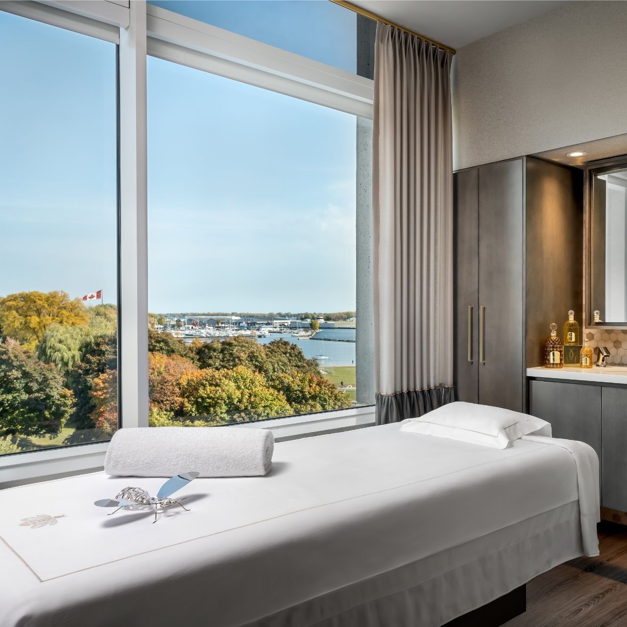 Spa treatment room with massage table and floor-to-ceiling windows overlooking waterfront and autumn foliage 