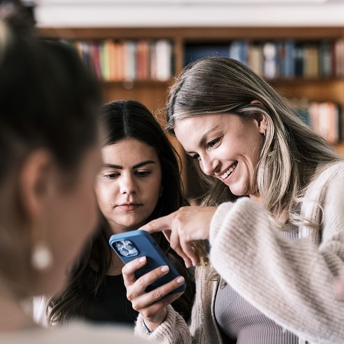 Three women discussing something on a phone in front of bookshelves at Falkensteiner Club Funimation Katschberg