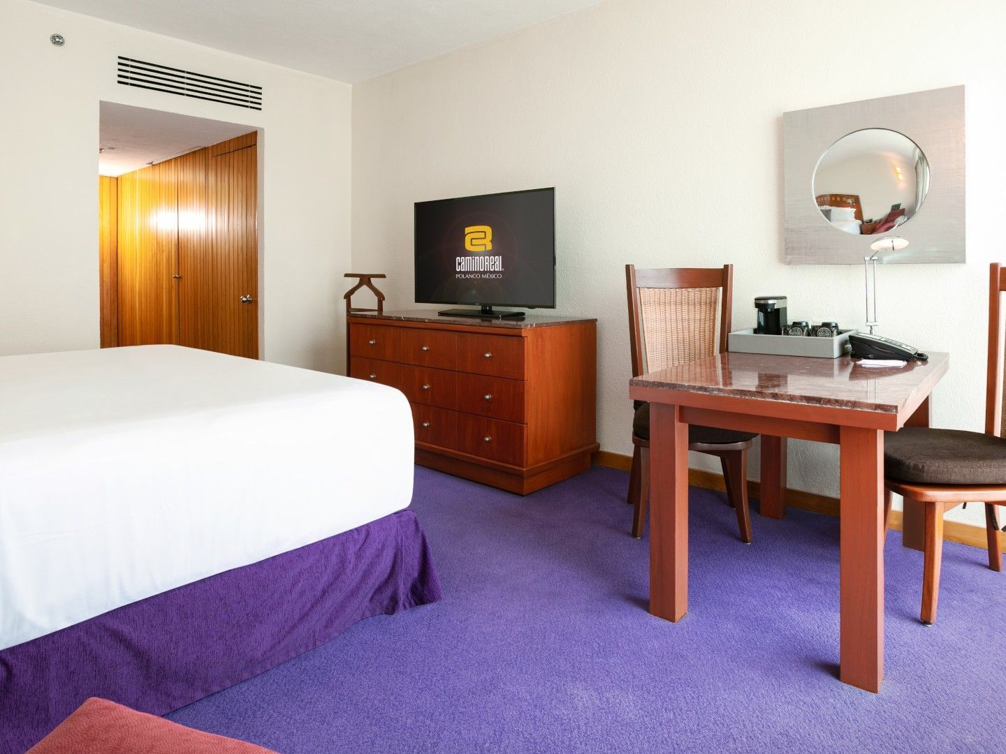 Habitación de hotel con cama grande, mesa, sillas, televisor y mobiliario en tonos cálidos.