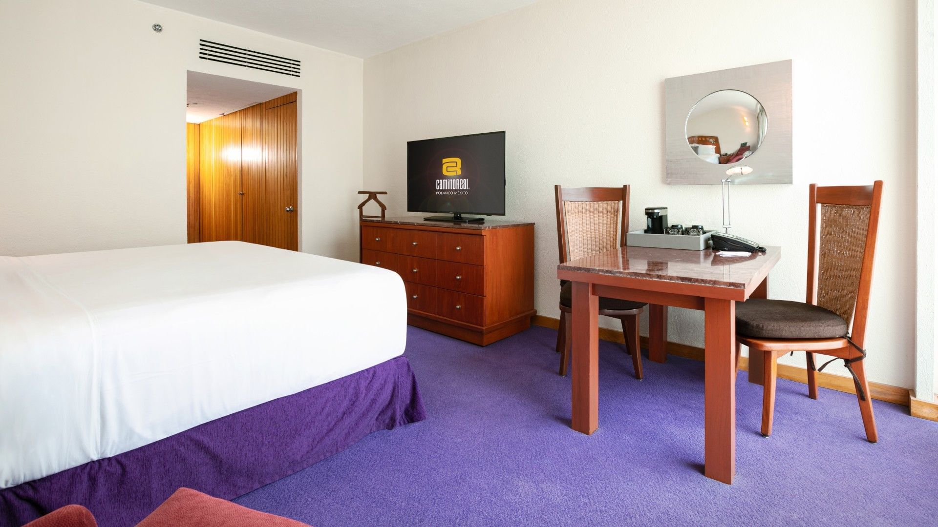 Habitación de hotel con cama grande, mesa, sillas, televisor y mobiliario en tonos cálidos.