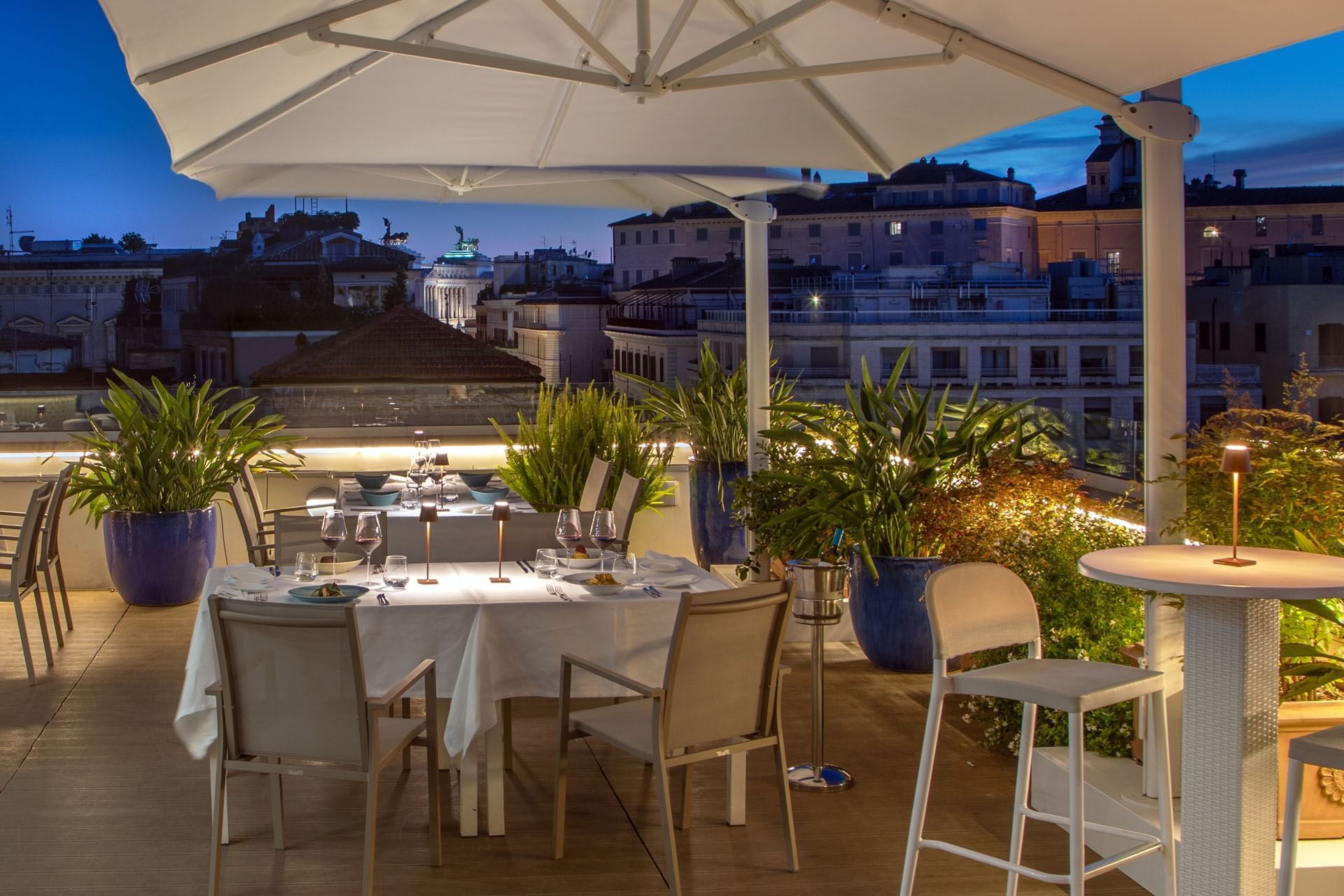 Elegant rooftop dining setup with wine glasses and lush greenery, overlooking city rooftops at The Tribune Hotel