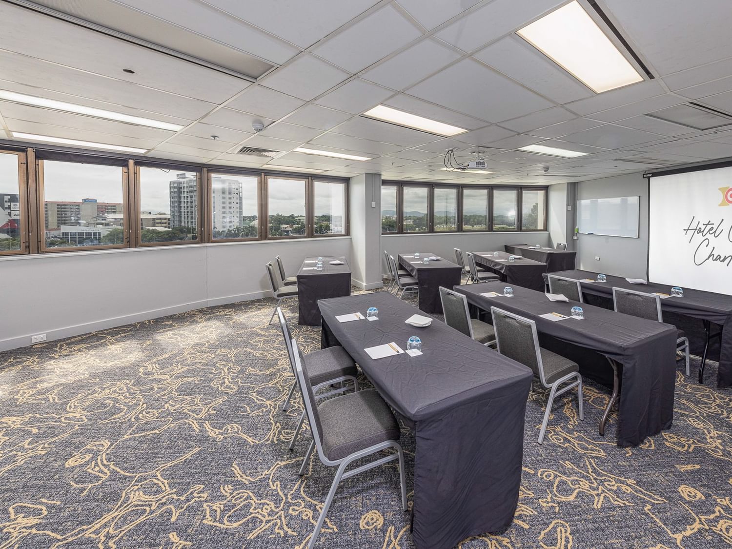 Classroom style conference setup in the Ross Creek Room, featuring black-clothed tables, grey chairs, windows, and a projector screen.