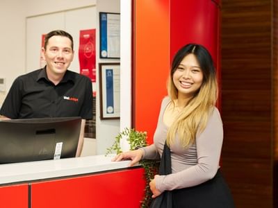 A smiling resident stands at a reception desk with a smiling staff member behind it.