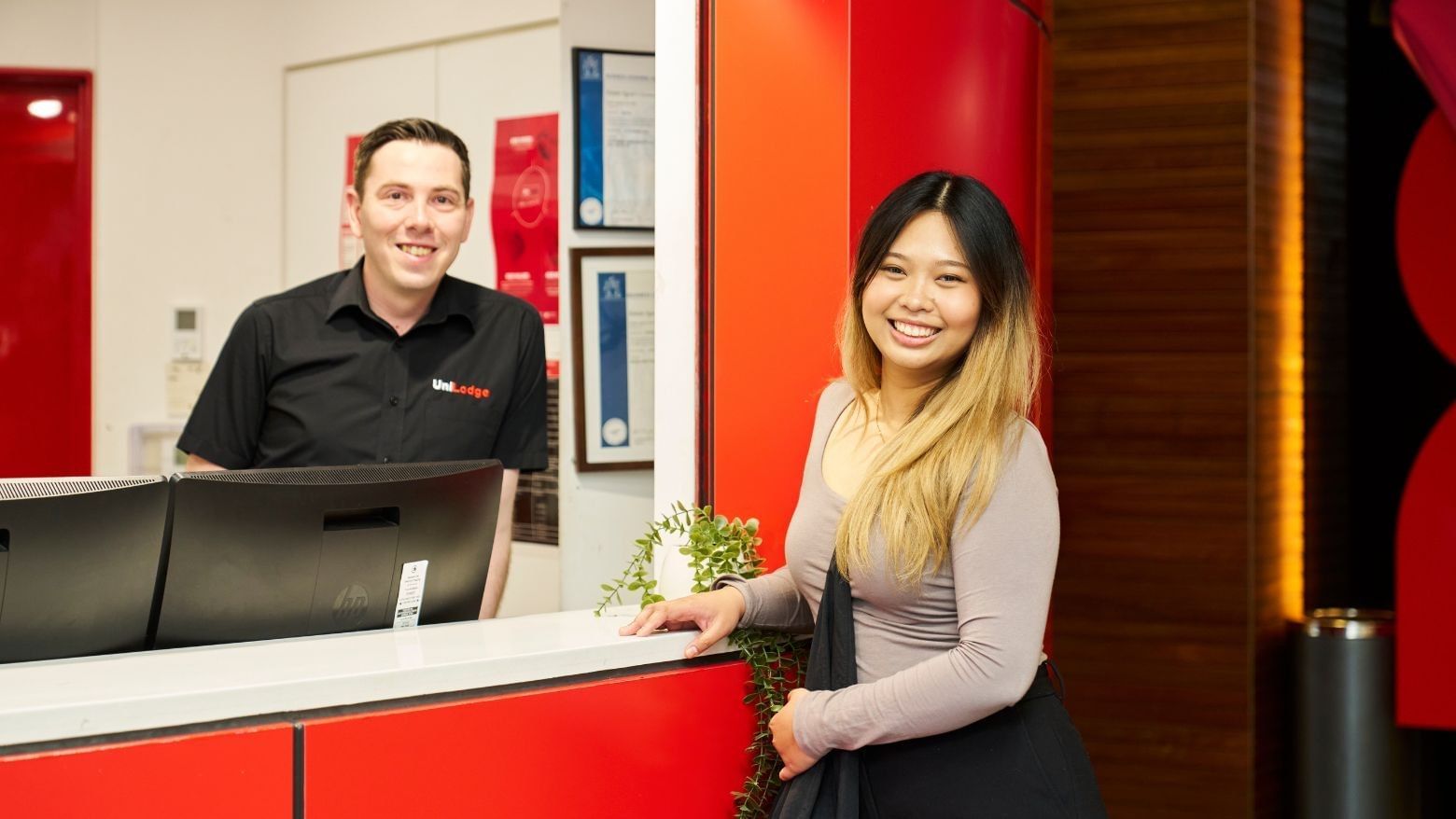 A smiling resident stands at a reception desk with a smiling staff member behind it.