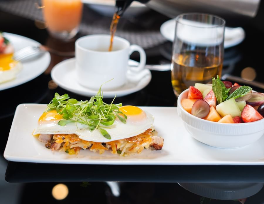 Fried eggs over hash browns by a fresh fruit bowl under a coffee pour at Warwick Melrose Dallas