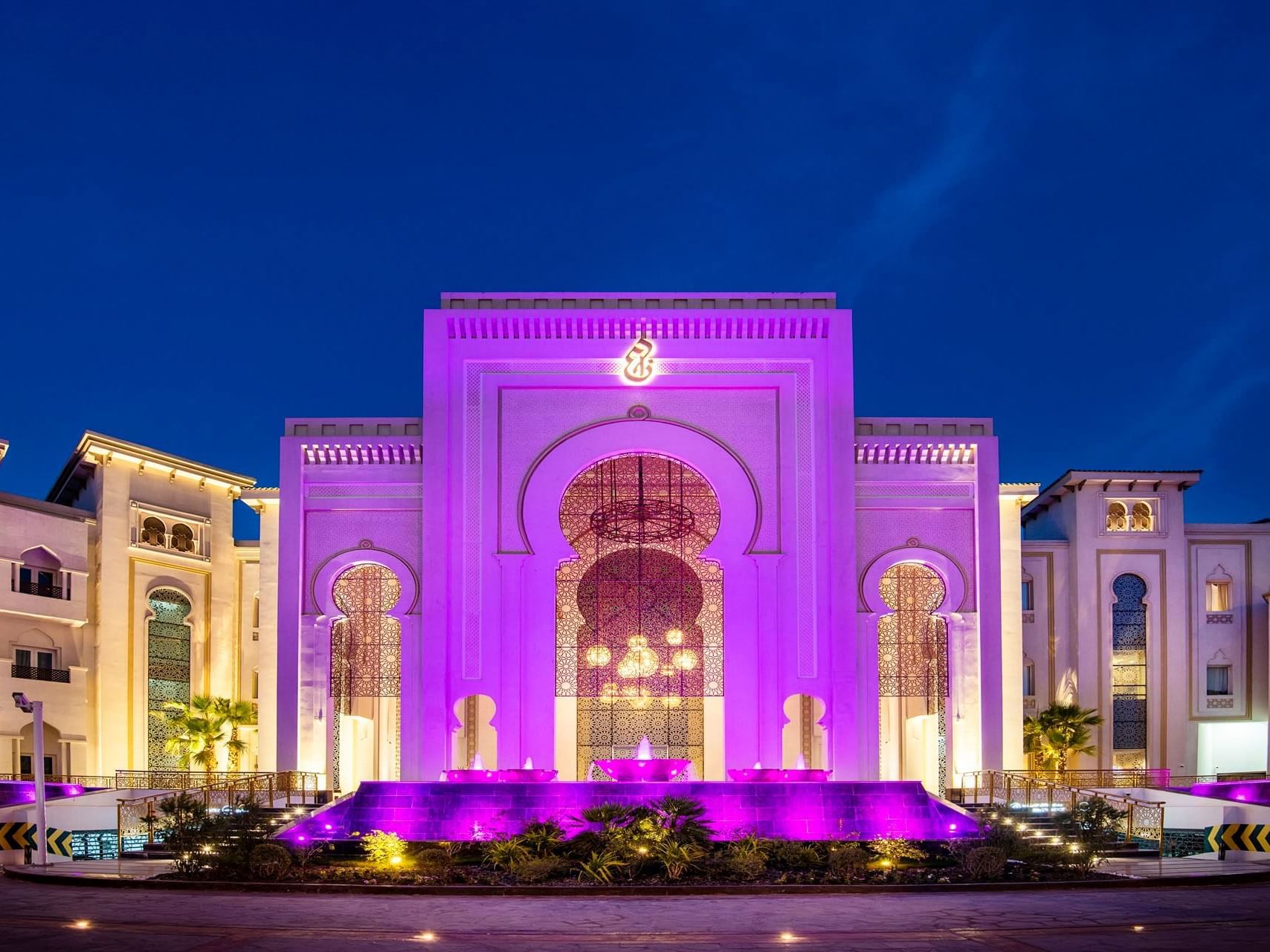 Illuminated ornate building at dusk with a large central arch and surrounding purple lighting.