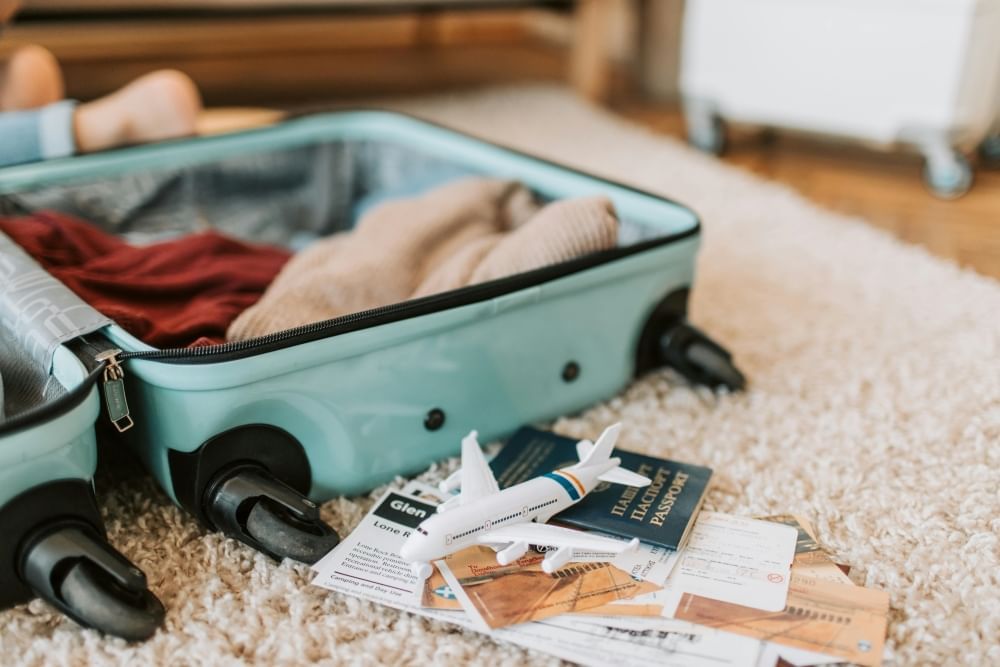 Half of an open light blue suitcase resting on white shag carpet with a few clothing items inside and a stack of tickets and paper next to it with a toy plane on top. 