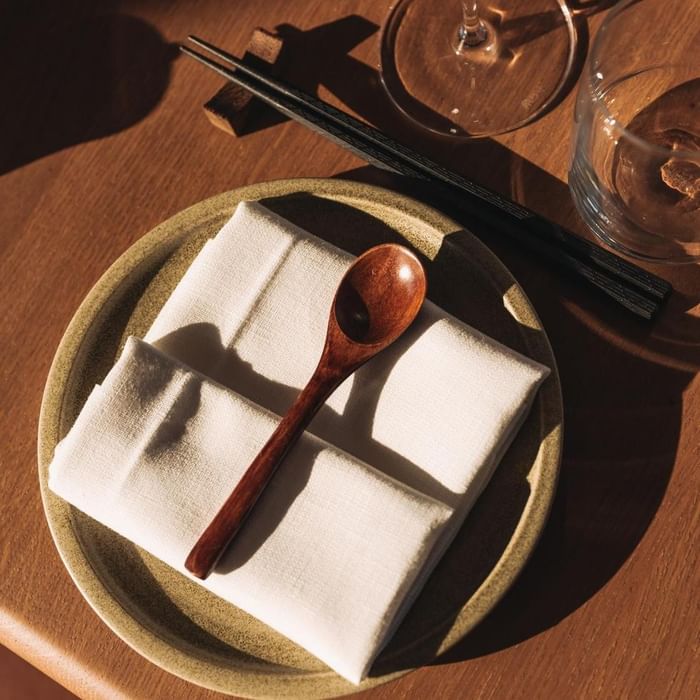 Holzlöffel auf Serviette, daneben Essstäbchen und ein Weinglas auf einem Tisch.