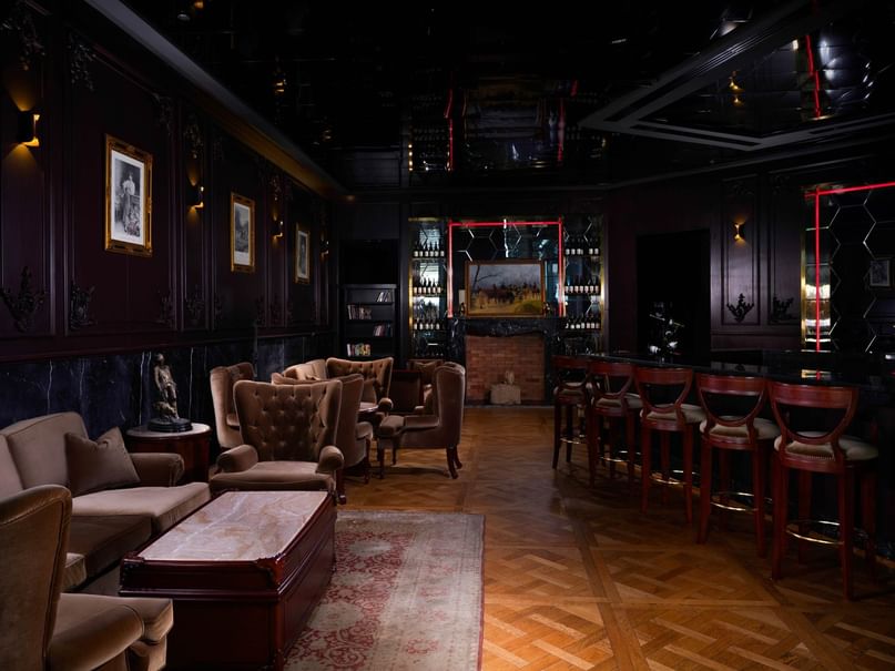 Dark bar interior with couches, chairs, and a bar counter, illuminated by dim lighting and wall-mounted frames.