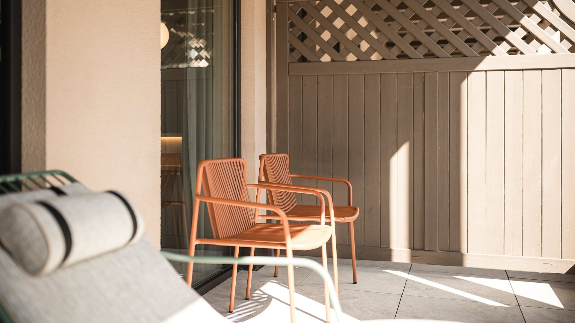 Balkon mit zwei orangefarbenen Stühlen, einer grünen Liege und einem Lattenzaun im Hintergrund.