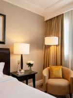 Tan armchair by a dark side table under a floor lamp near a white bed in The Montaigne Suite at Warwick Paris Champs Elysées