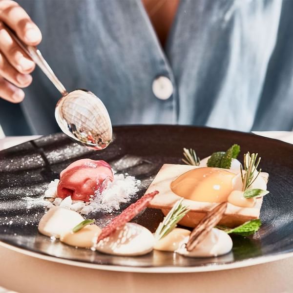 Hand holding a spoon adds a sauce to an elegantly plated dessert at Falkensteiner Hotels & Residences