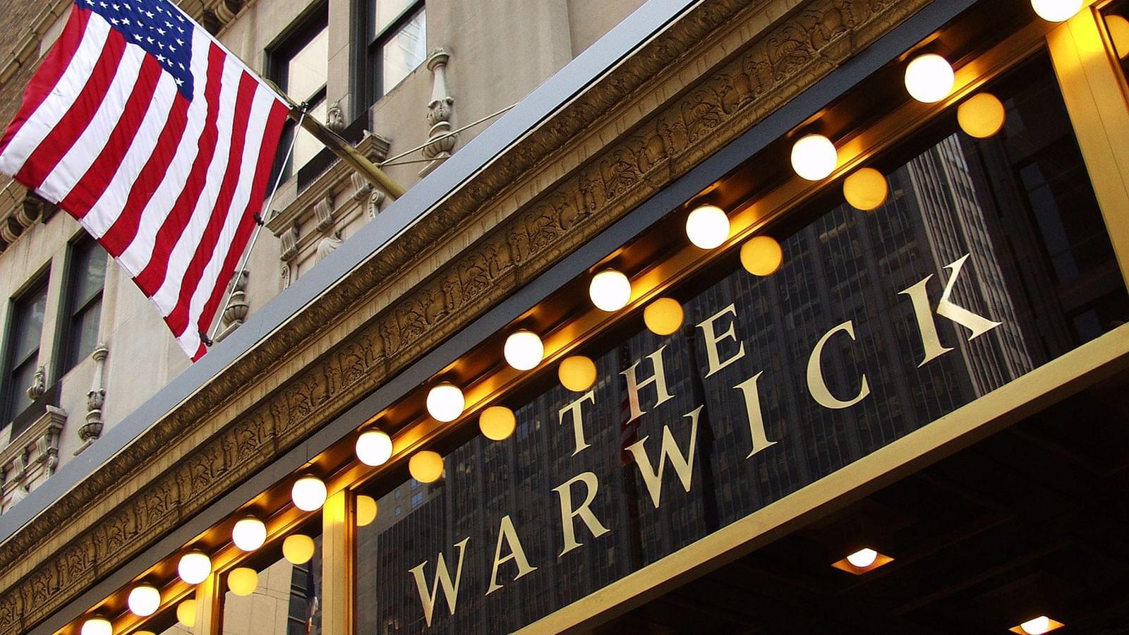 Golden illuminated marquee sign for The Warwick hotel with an American flag above it