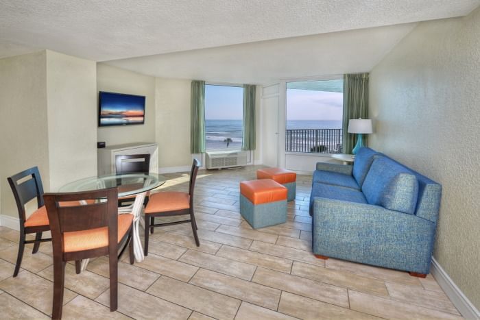 A wide view of a hotel suite with a blue sofa, glass dining table, fireplace, and ocean view.