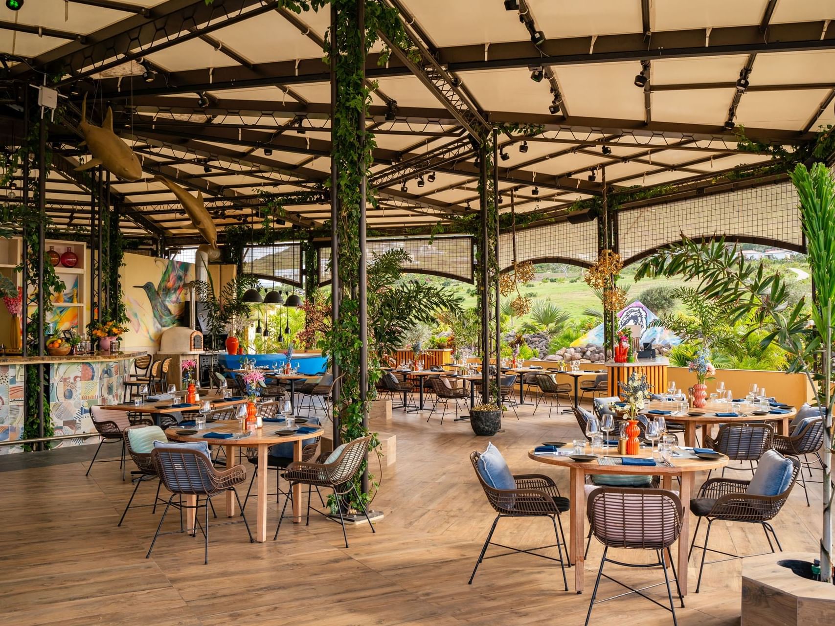 Eetgedeelte met tafels en gevlochten stoelen, gevuld met planten en decoratie in Bobbie’s Beach Club bij Golden Rock Resort