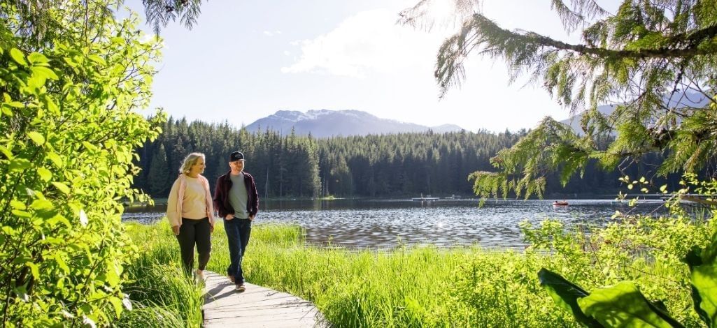 Couple walks along a wooden path near Lost Lake in Whistler surrounded by trees, water and mountain scenery