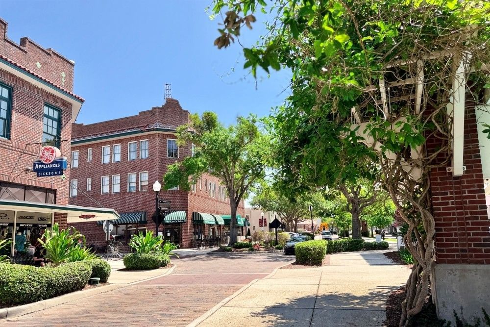 A cobblestone street with brick buildings, mature trees, climbing vines, and shops with green awnings. 