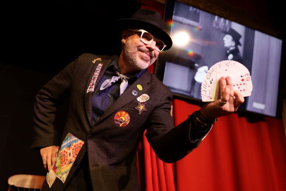 A man in a suit and glasses holds a fan of playing cards while smiling onstage.