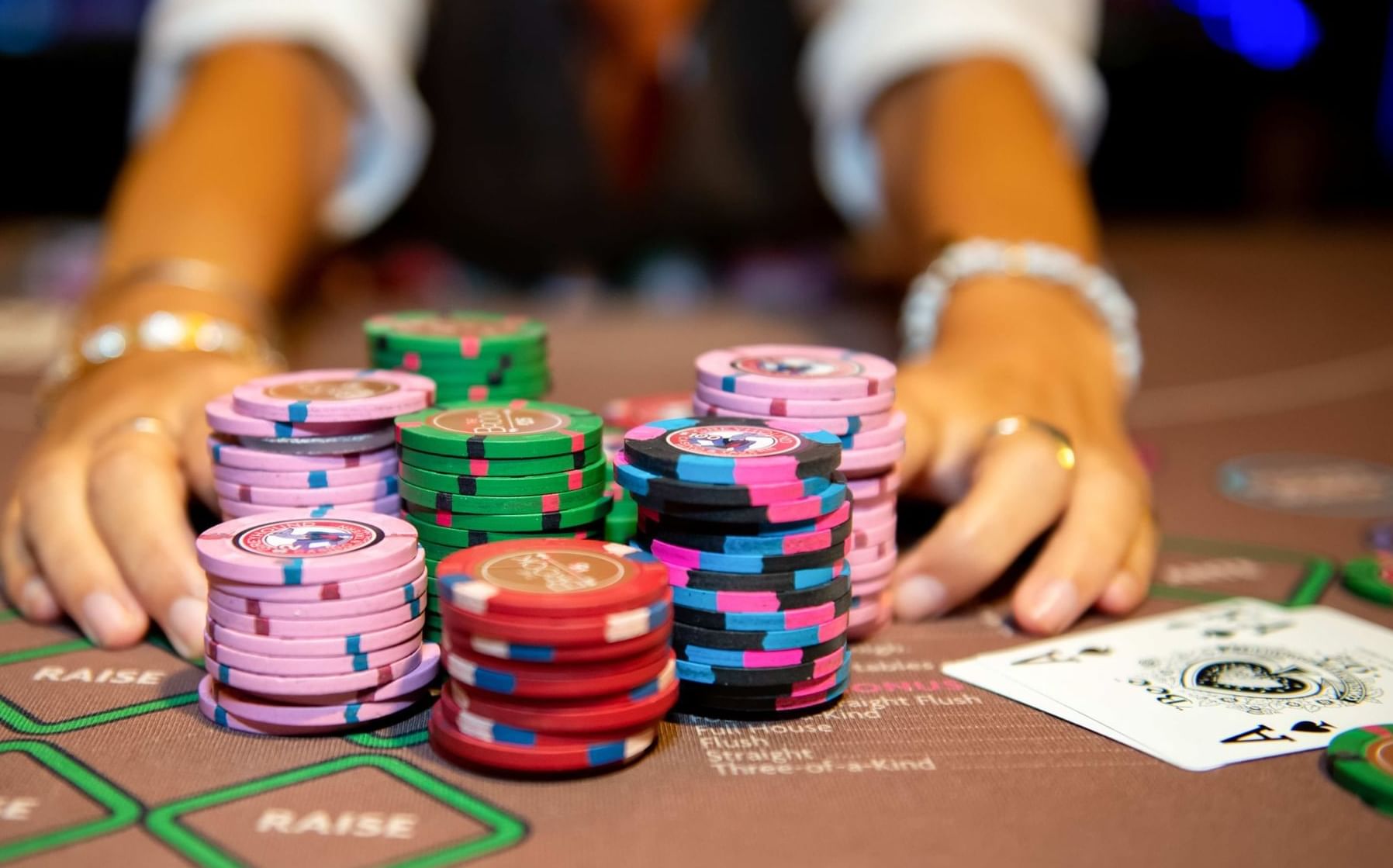 Colorful casino chips stacked by playing cards on a gaming table as a dealer reaches forward inside The Brook