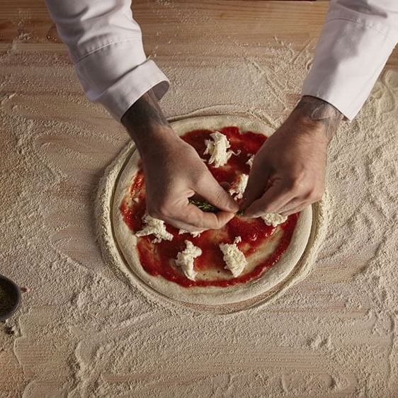 Chef's hands sprinkle cheese on a tomato-sauced pizza dough in restaurant at AG Hotels