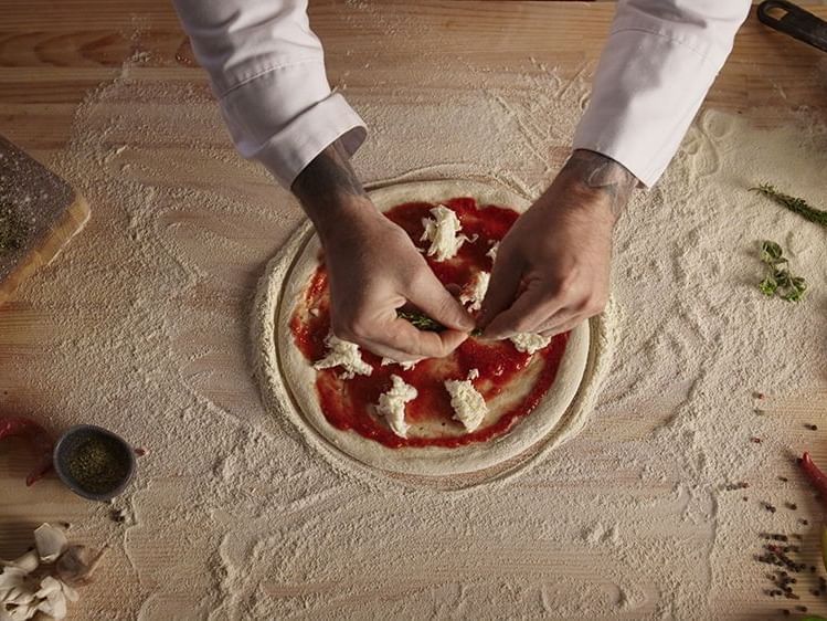 Chef's hands sprinkle cheese on a tomato-sauced pizza dough in restaurant at AG Hotels