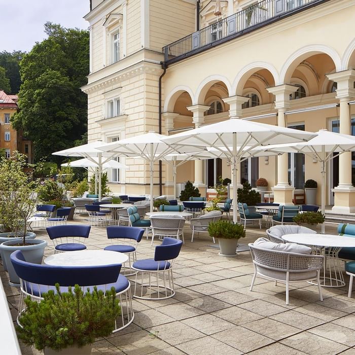 Eine elegante Terrasse mit weißen Sonnenschirmen, blauen Stühlen und Tischen vor einem Gebäude mit Säulen.