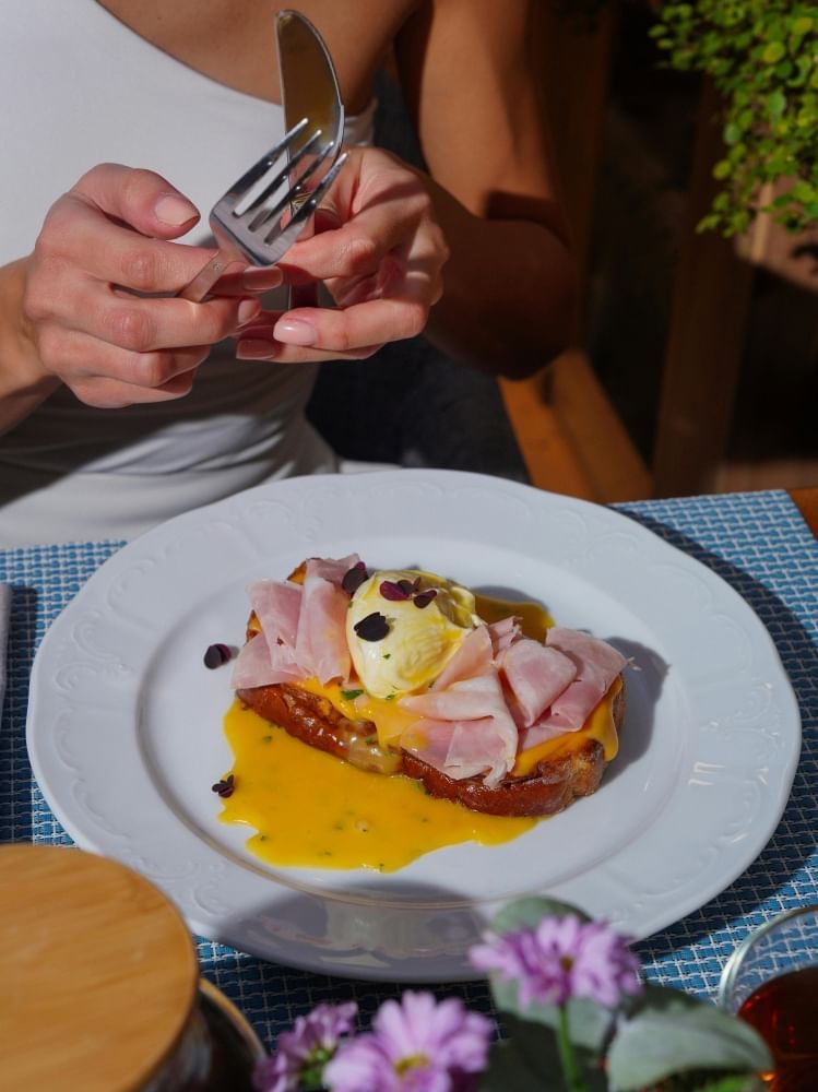 A close-up of hands and a white plate with a slice of toast topped with ham, a poached egg, and garnishes. 