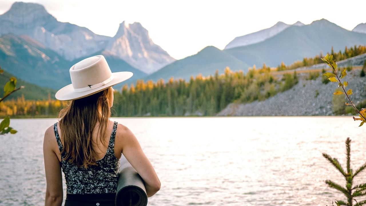Woman in a hat stands by a lake with mountains in the background.