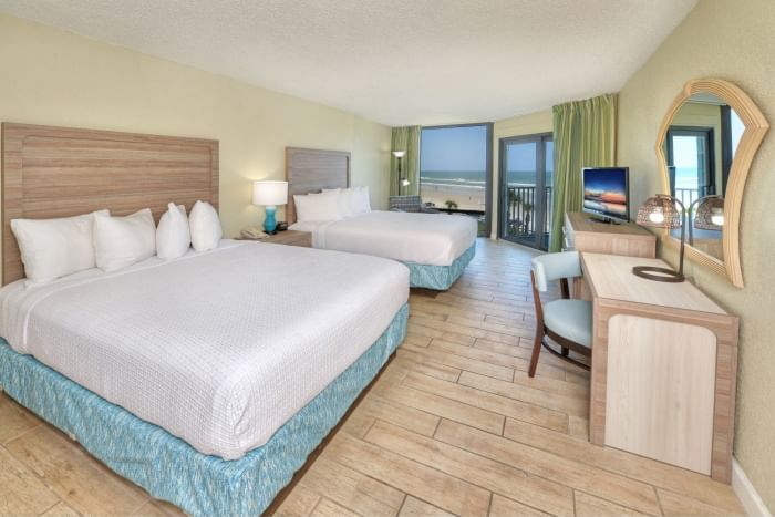 A wide view of a hotel room with two beds, a desk, a TV, and a balcony with a beach view.
