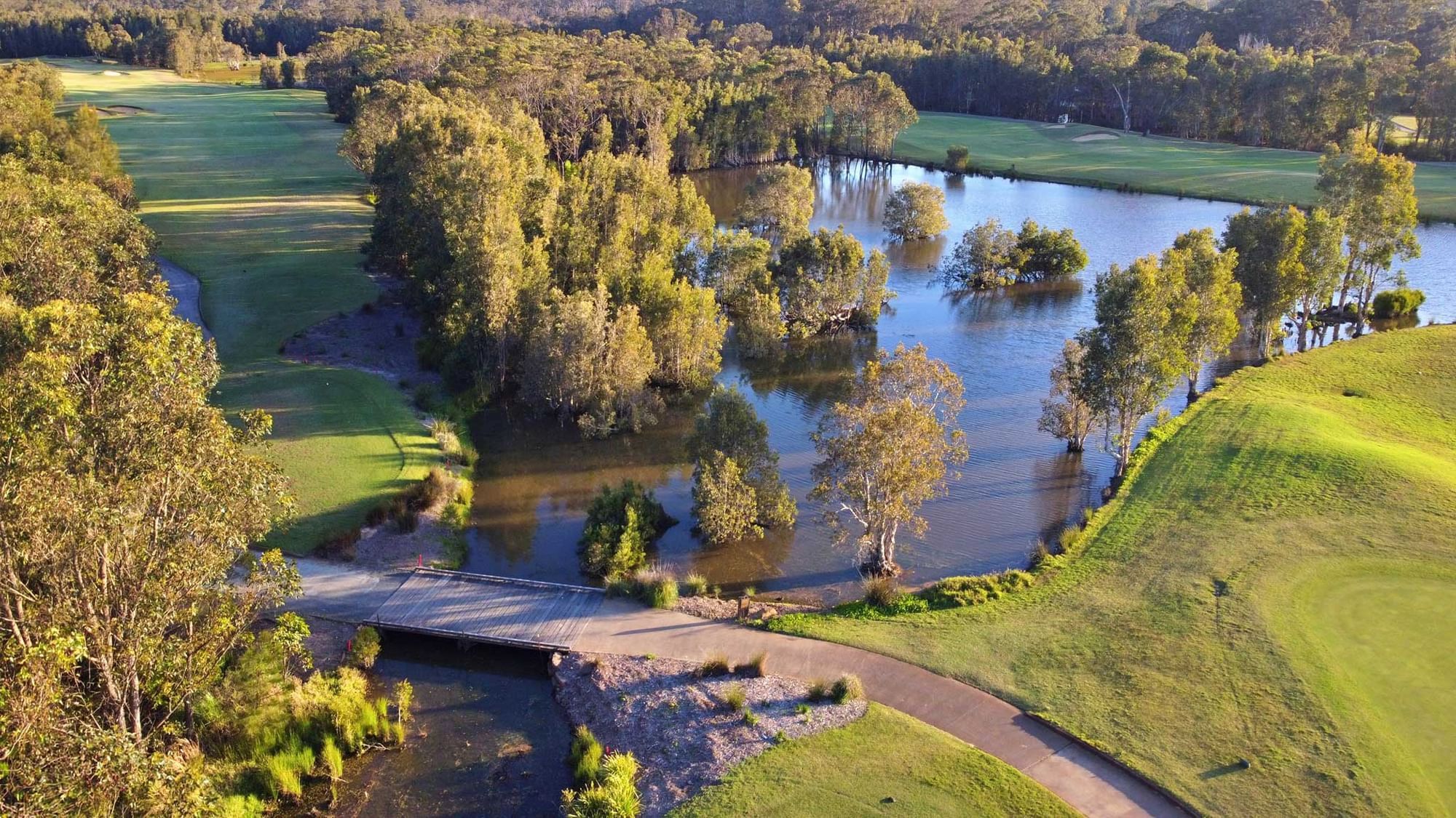 Aerial view of a golf course with a flooded area, bridge, and trees surrounding the green field.