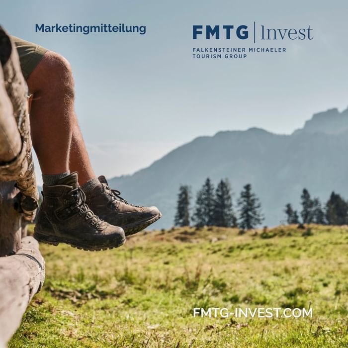 Füße auf Holzbalken über Wiese mit FMTG Invest Logo und Berglandschaft im Hintergrund.