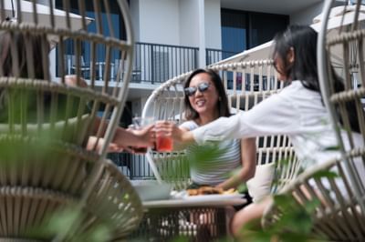 Three women clinking glasses in wicker chairs on a patio with sun loungers and tables at the Maui Coast Hotel