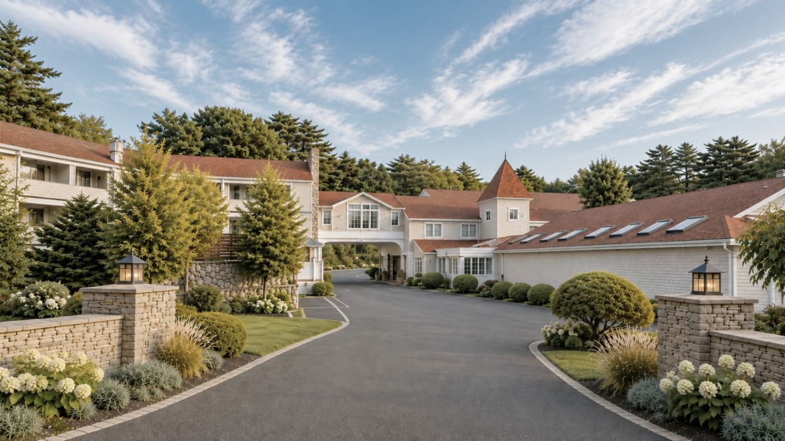 A large white building with a brown roof and a paved driveway surrounded by trees and stone walls.