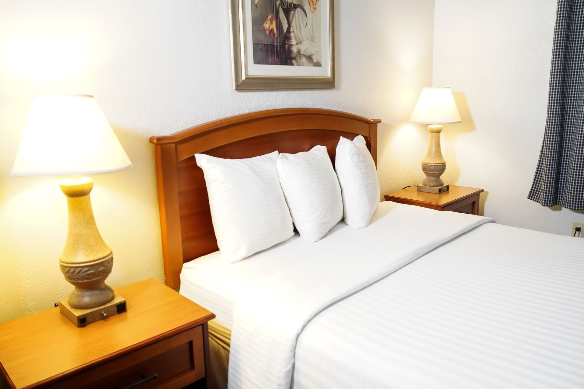 A neatly made hotel room with a wooden bed frame, white bedding, two nightstands with lamps, and a framed picture on the wall.