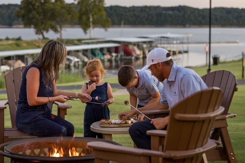 Family roasts marshmallows over a fire pit in adirondack chairs by a lakeside dock at Shangri-La Resort and Golf Club