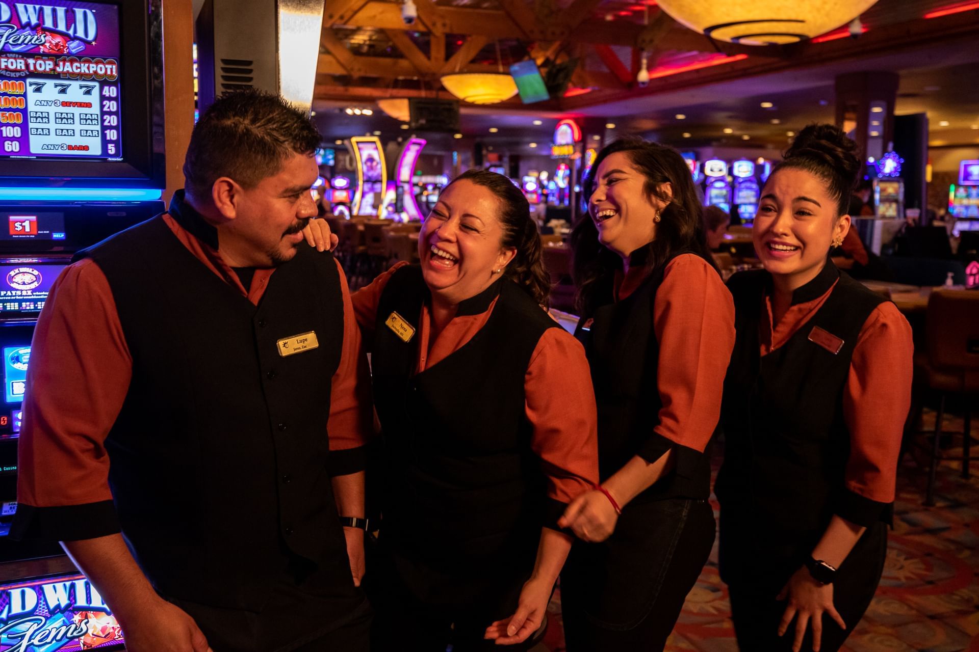 Four casino employees wearing matching uniforms are laughing together near slot machines inside Eureka Casino Resort