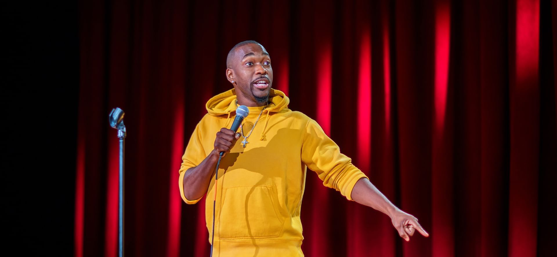 Comedian Jay Pharoah wearing yellow hoodie performing live on stage with red curtain backdrop venue at The Brook