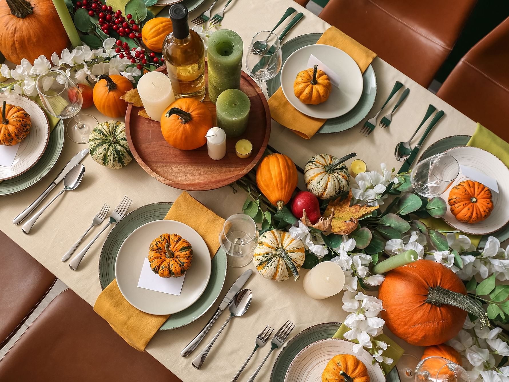 A table with place settings, pumpkins, candles, flowers, and a bottle of wine, surrounded by chairs.