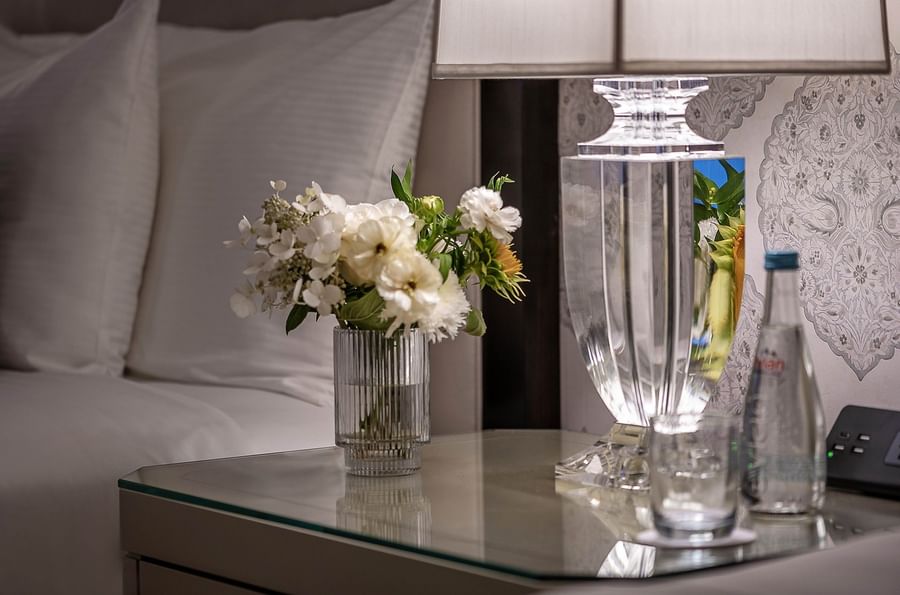 Flowers, lamp, and bottle on a glass table next to a bed.