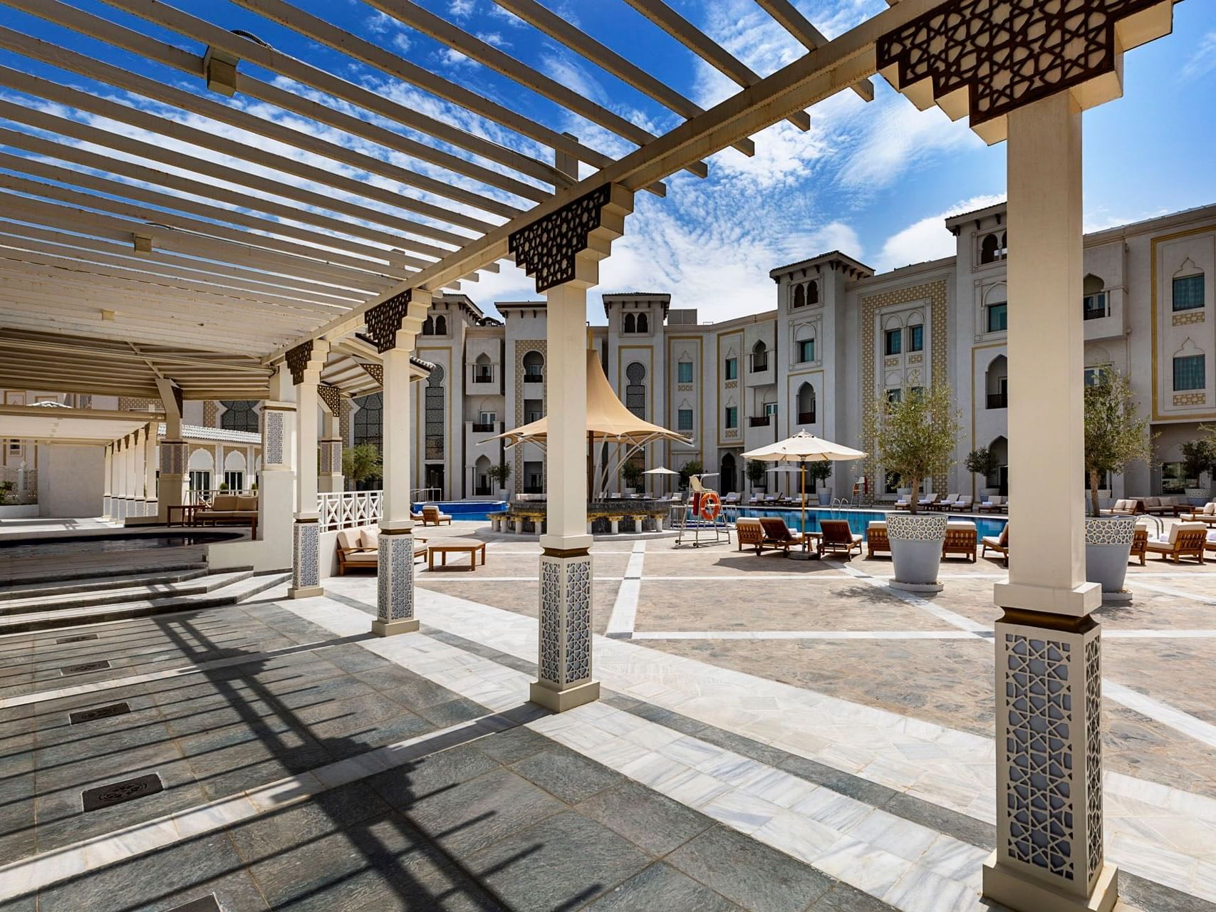Covered outdoor patio area with lounge chairs and a pool in the background under a blue sky.