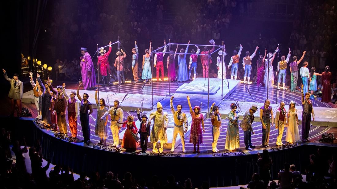 Performers in costumes on a circular stage with raised hands, surrounded by an audience in a dark venue.